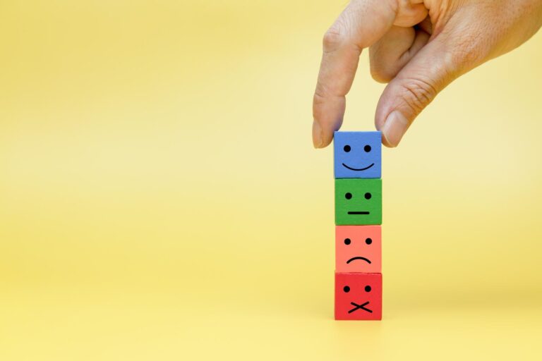 A person's hand stacking blocks with facial expressions drawn on them. The bottom block is angry, the next is disappointed, the next is indifferent and the last block is happy