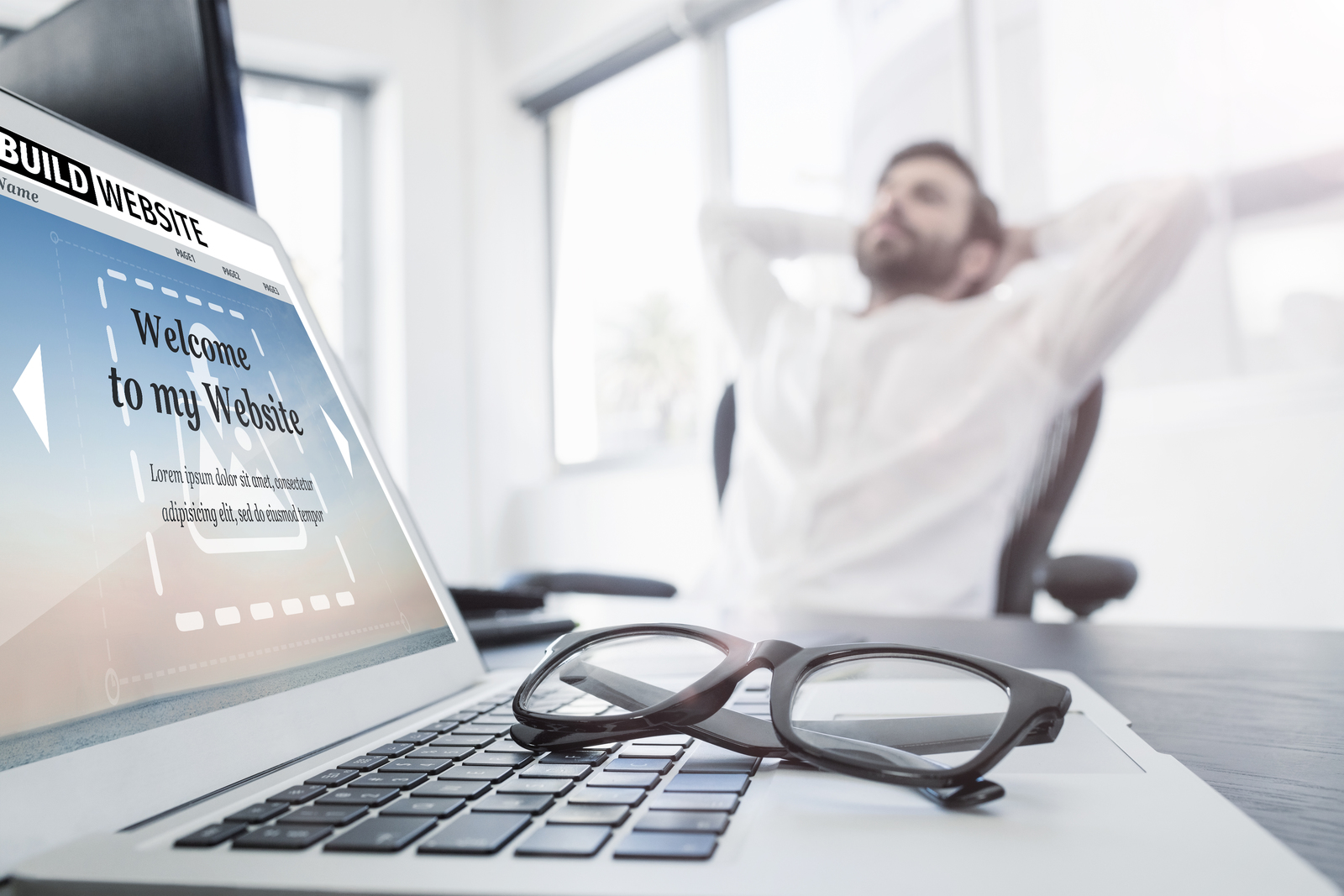 Man sitting back thinking in a chair with an open laptop displaying image of a new website without content. His glasses are folded on top of the keyboard.