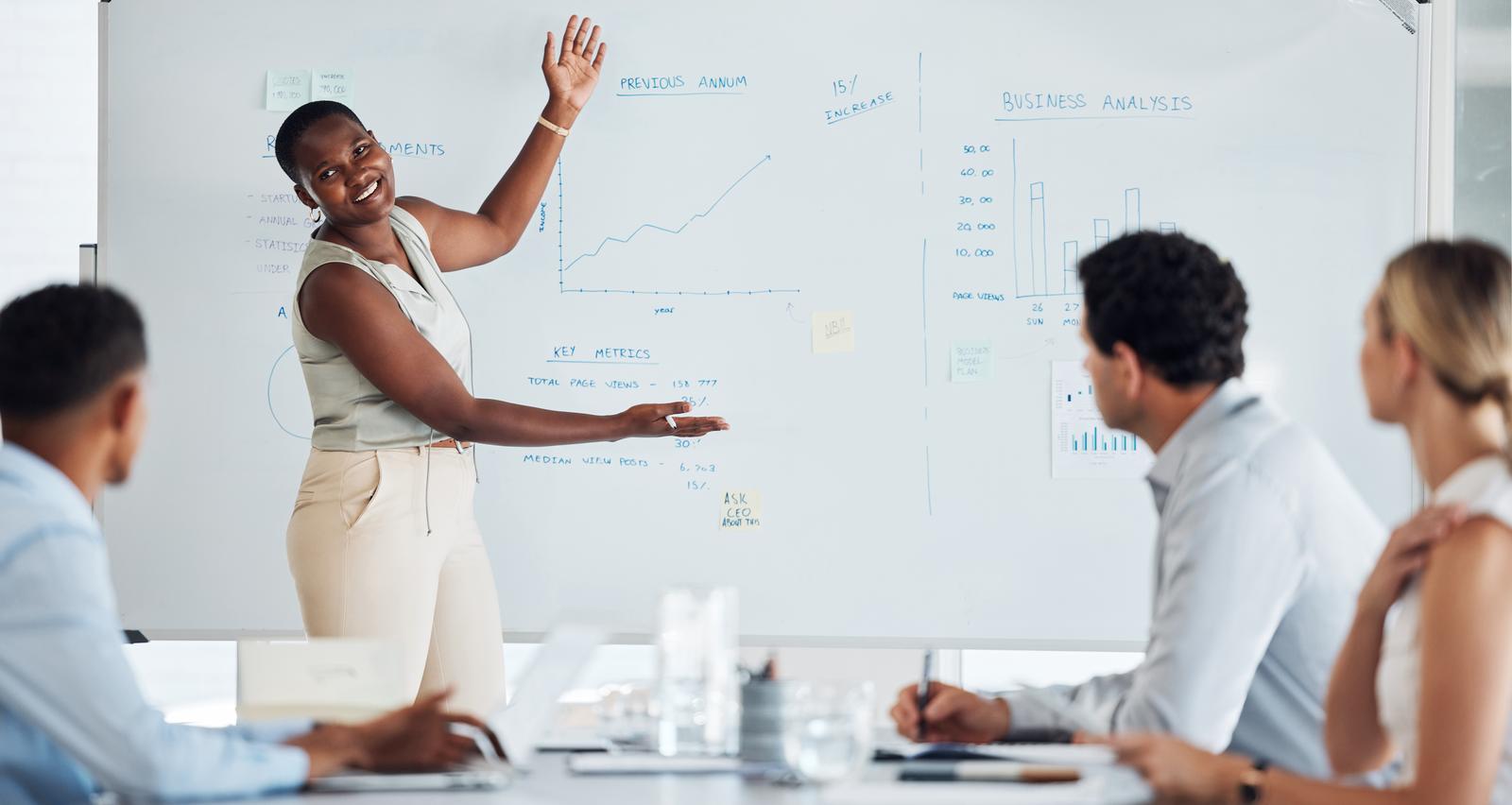 woman standing infront of a whiteboard with key metrics and other data written on the board. She is presenting the data to two men and a woman sat around a table taking notes.