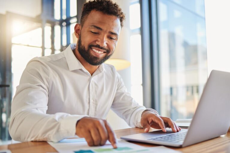 Business man sat at a desk carrying out web scraping using his laptop while pointing to data on a print out.