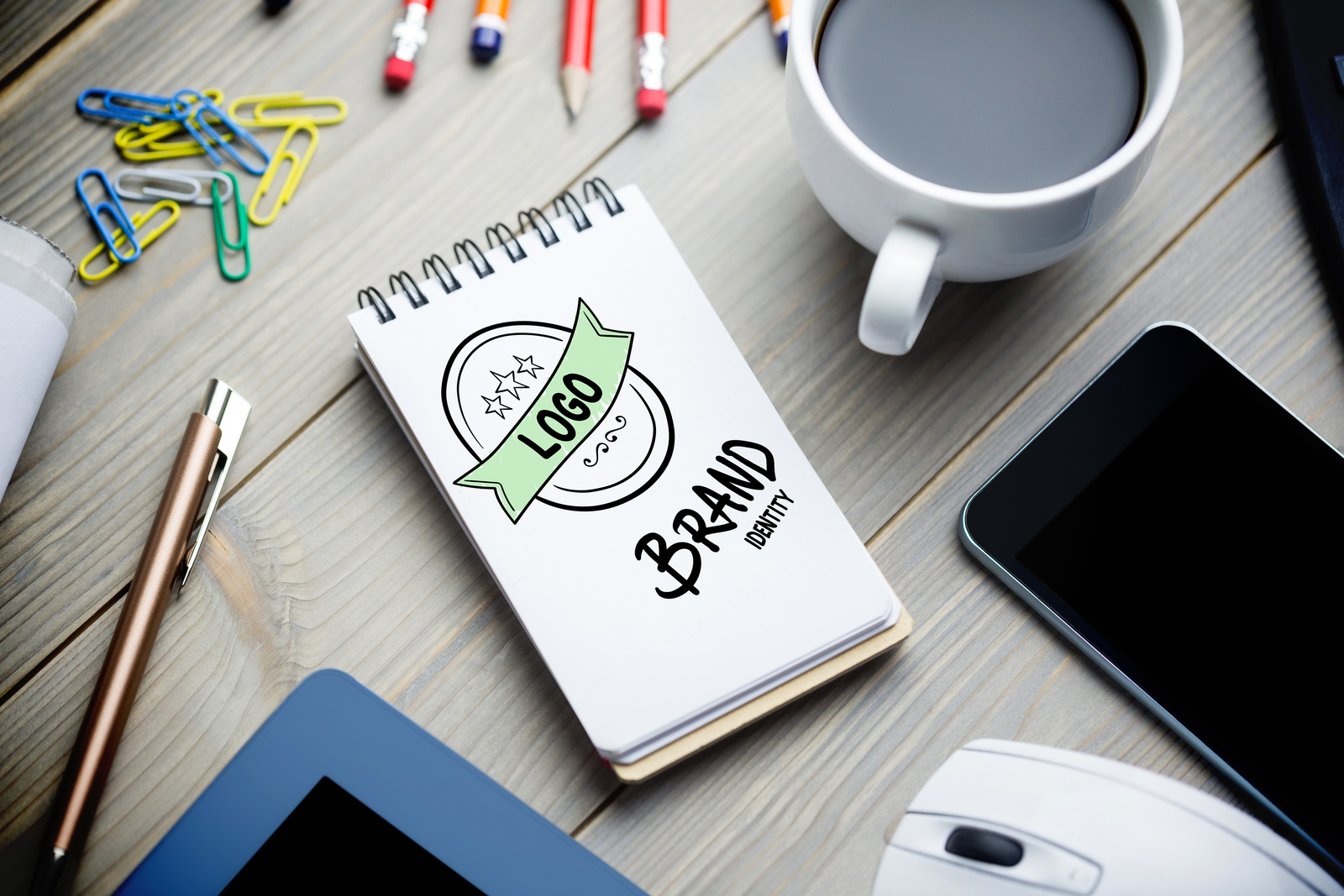 Table with a cup of coffee, pens, computer mouse, smartphone and pad with a logo and the words Brand Identity at the bottom.