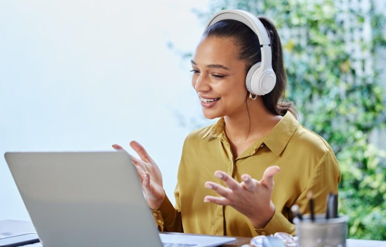 Young woman wearing headphones, sat in front of a laptop with her hands in front of her smiling