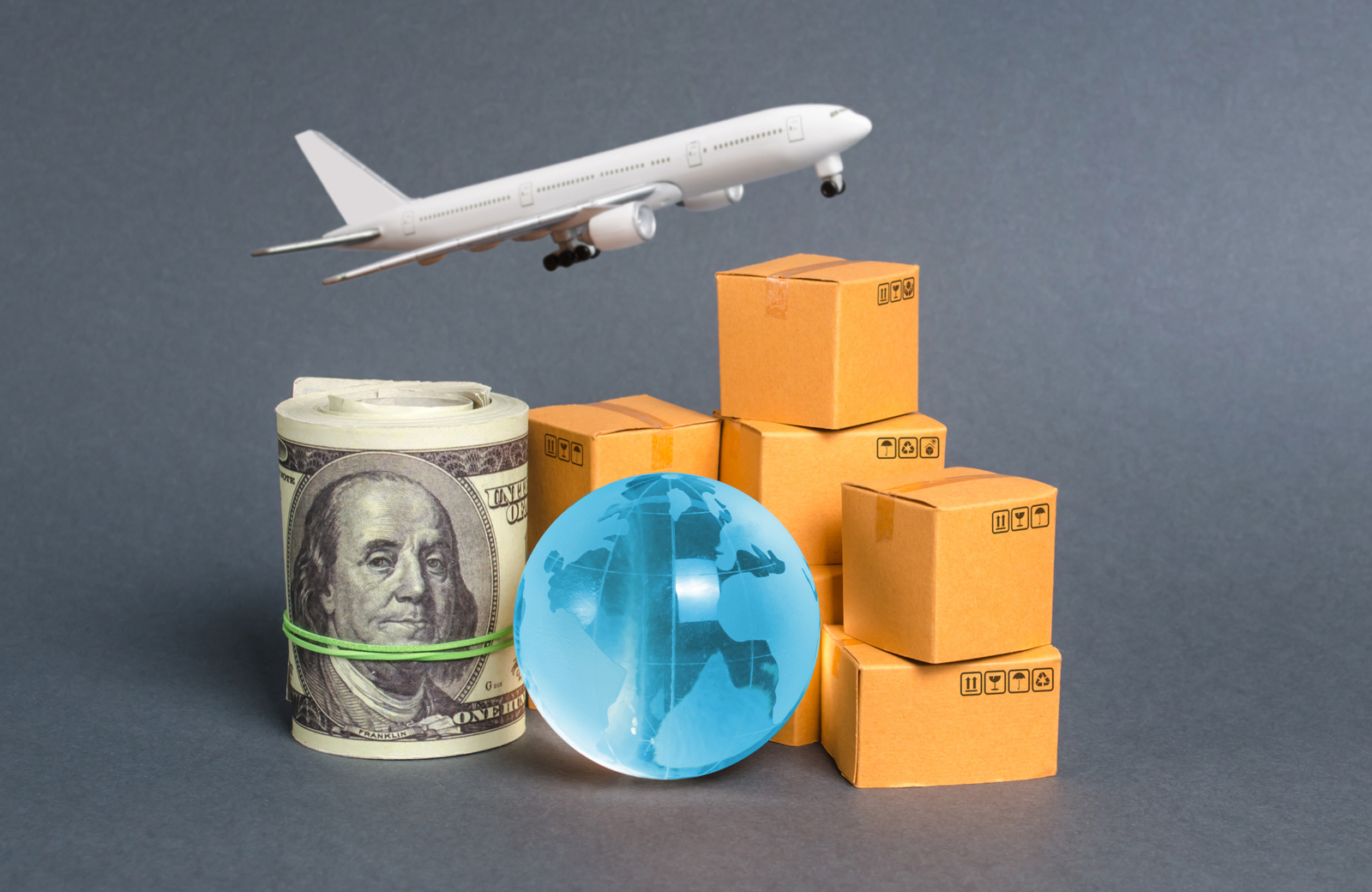 A plane, roll of dollar notes, a globe and boxes for export against a grey background